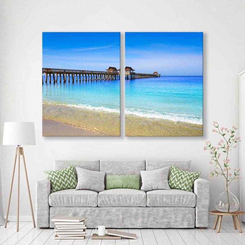 May include: A split canvas print of a wooden pier extending into a turquoise ocean with a sandy beach in the foreground. The sky is a bright blue with white clouds.