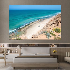 May include: A large canvas print of a coastal scene. The image features a vibrant turquoise ocean meeting a sandy beach, with a rocky cliffside and clear blue sky. The artwork is displayed above a bed in a modern bedroom.