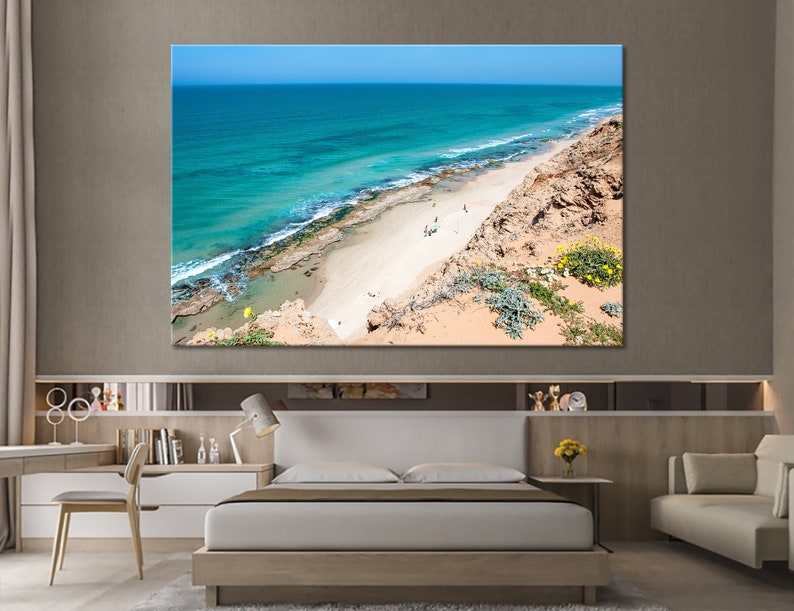 May include: A large canvas print of a coastal scene. The image features a vibrant turquoise ocean meeting a sandy beach, with a rocky cliffside and clear blue sky. The artwork is displayed above a bed in a modern bedroom.