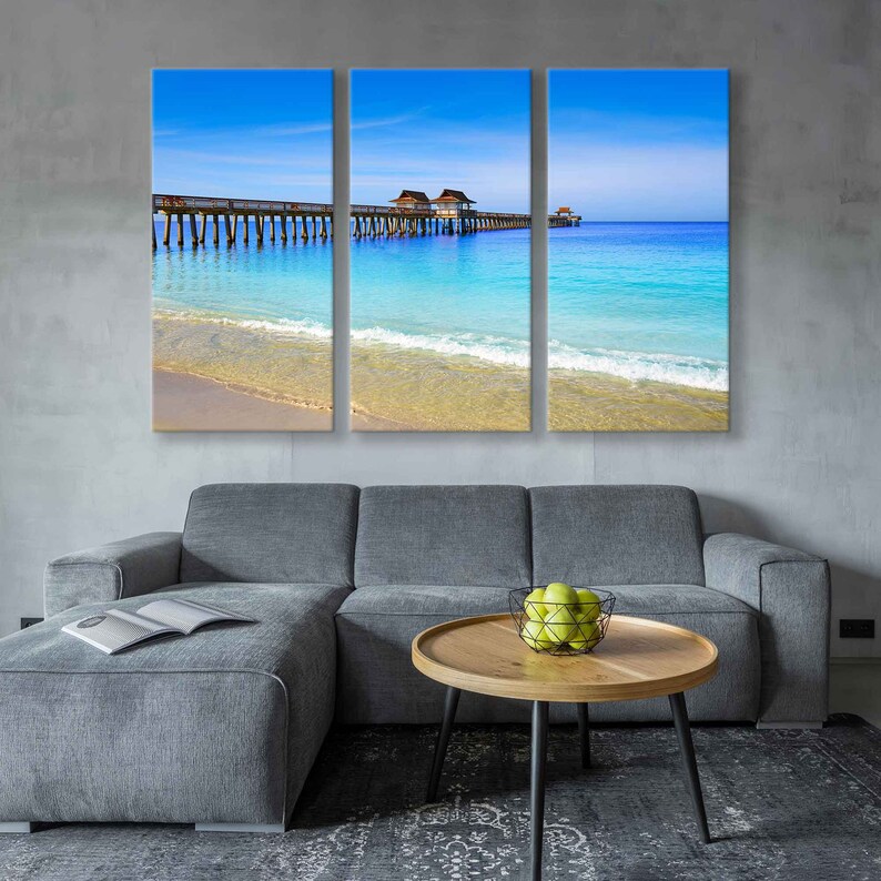 May include: A three-panel canvas print featuring a wooden pier extending over a turquoise ocean with white waves crashing on the sandy shore. The sky is a bright blue with white clouds.