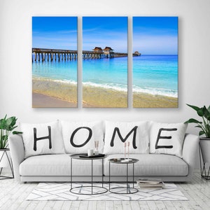 May include: A three-panel canvas print featuring a beach scene with a wooden pier extending into the turquoise water. The sky is a bright blue with fluffy white clouds. The word "HOME" is written in black on white pillows on a white couch in the foreground.