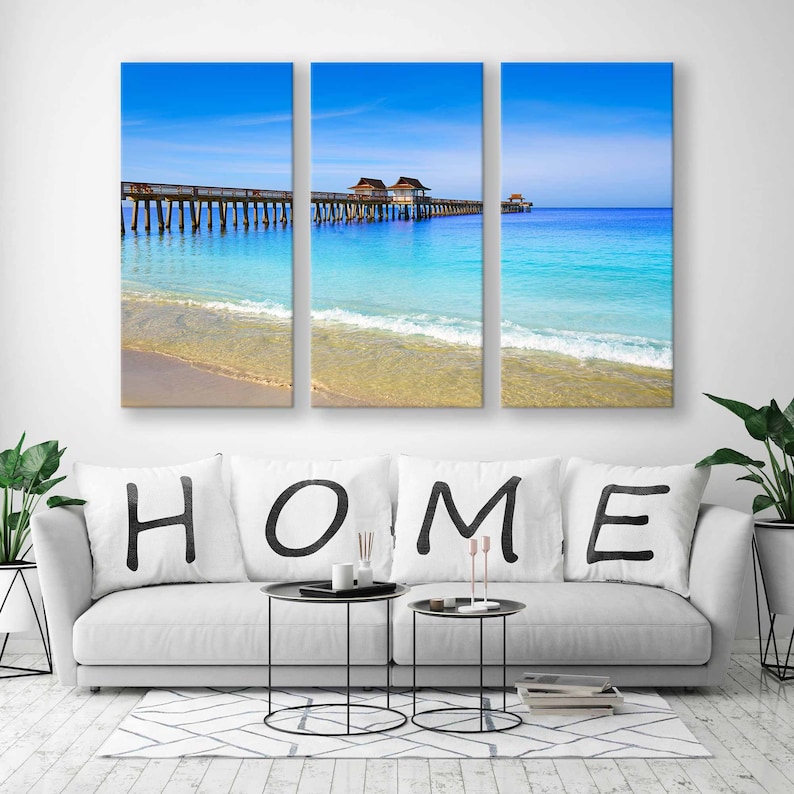May include: A three-panel canvas print featuring a beach scene with a wooden pier extending into the turquoise water. The sky is a bright blue with fluffy white clouds. The word "HOME" is written in black on white pillows on a white couch in the foreground.