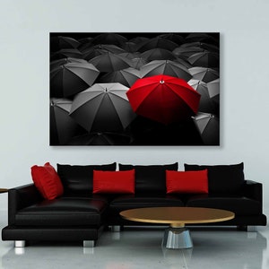May include: A canvas print featuring a striking contrast of umbrellas. The majority are black and gray, with one vibrant red umbrella standing out. The artwork is displayed above a black sectional sofa with red throw pillows and a round coffee table.