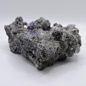 May include: A raw mineral specimen featuring a dark, porous, and textured grey matrix embedded with clusters of translucent, cubic purple and clear crystals. The geological formation has an irregular, jagged shape with visible crystalline structures.