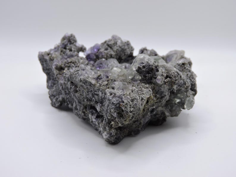 May include: A raw mineral specimen featuring a dark, porous, and textured grey matrix embedded with clusters of translucent, cubic purple and clear crystals. The geological formation has an irregular, jagged shape with visible crystalline structures.