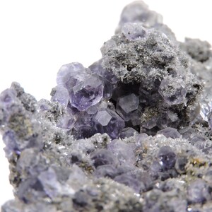 May include: A cluster of translucent purple fluorite crystals with geometric cubic formations embedded in a rough, grey, textured mineral matrix. The crystal surfaces exhibit sharp edges and varying shades of violet, contrasting with the dark, granular rock base.