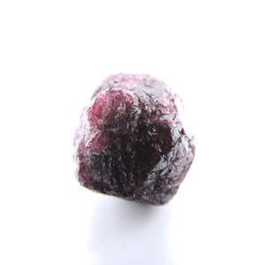 May include: A raw, unpolished garnet gemstone with a deep, dark red and purple hue. The stone features a naturally irregular, multifaceted surface with visible crystalline textures and a semi-translucent quality throughout its dense structure.