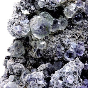 May include: A mineral specimen featuring clusters of translucent, purple-tinted cubic crystals embedded within a dark, rough, and granular matrix. The geometric crystal formations vary in size, displaying sharp edges and a glassy, reflective surface.