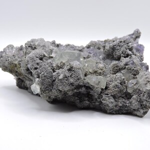 May include: A raw mineral specimen featuring a cluster of translucent, pale purple and clear cubic crystals embedded within a rough, dark grey, porous volcanic rock matrix. The crystalline structure displays sharp geometric edges and facets.