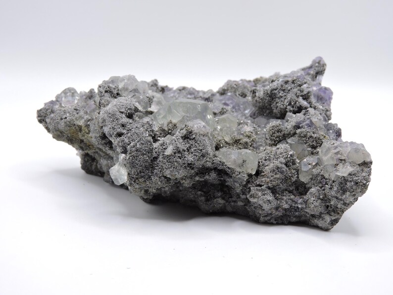 May include: A raw mineral specimen featuring a cluster of translucent, pale purple and clear cubic crystals embedded within a rough, dark grey, porous volcanic rock matrix. The crystalline structure displays sharp geometric edges and facets.