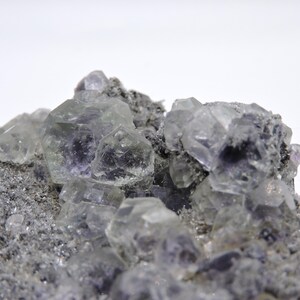 May include: A cluster of translucent, pale purple fluorite crystals with geometric, cubic facets embedded in a rough, grey, granular mineral matrix. The crystalline structures exhibit varying degrees of clarity and light reflection across the surface.