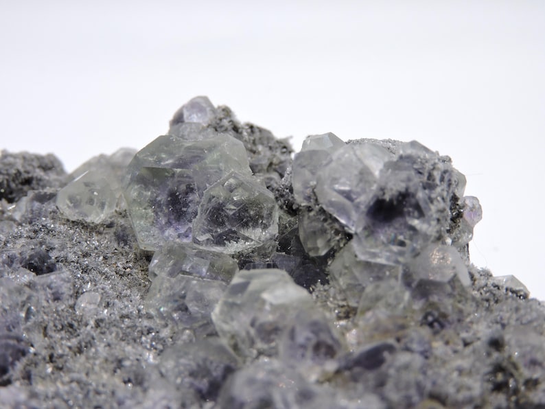 May include: A cluster of translucent, pale purple fluorite crystals with geometric, cubic facets embedded in a rough, grey, granular mineral matrix. The crystalline structures exhibit varying degrees of clarity and light reflection across the surface.