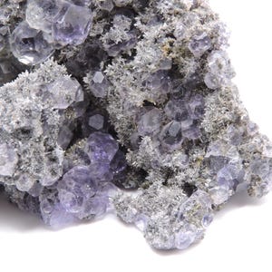 May include: A mineral specimen featuring clusters of translucent, light purple fluorite crystals embedded within a textured, grey, crystalline matrix. The formation displays numerous geometric crystal faces and jagged, rough edges throughout the piece.