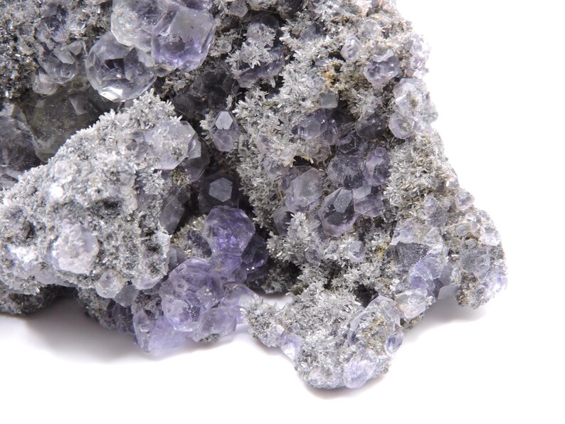 May include: A mineral specimen featuring clusters of translucent, light purple fluorite crystals embedded within a textured, grey, crystalline matrix. The formation displays numerous geometric crystal faces and jagged, rough edges throughout the piece.