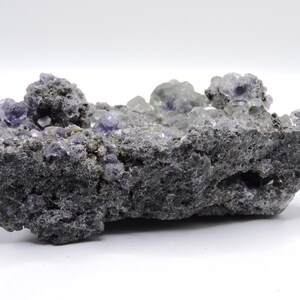 May include: A raw mineral specimen featuring clusters of translucent purple cubic crystals embedded within a dark, porous, and textured grey rock matrix. The crystalline formations are concentrated along the top edge, showcasing geometric shapes and varying shades of violet against the rough, jagged surface of the stone base.
