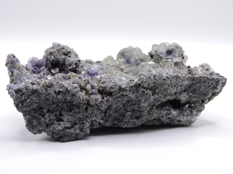 May include: A raw mineral specimen featuring clusters of translucent purple cubic crystals embedded within a dark, porous, and textured grey rock matrix. The crystalline formations are concentrated along the top edge, showcasing geometric shapes and varying shades of violet against the rough, jagged surface of the stone base.