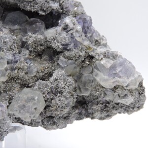 May include: A mineral specimen featuring translucent, pale purple cubic fluorite crystals embedded within a rough, dark grey, granular matrix. The cluster displays multiple geometric crystal formations with varying levels of clarity and light reflection.