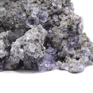 May include: A cluster of translucent purple fluorite crystals embedded in a rough, grey, granular matrix. The mineral specimen features numerous small, geometric cubic crystal formations with varying shades of violet and clear crystalline edges.