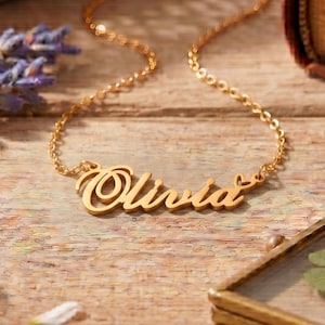 May include: A gold-toned metal necklace featuring a cursive nameplate pendant that reads Olivia, with a small heart detail at the end of the name. The pendant is attached to a delicate, thin gold-toned chain with a classic link design.