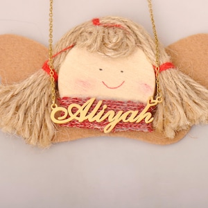 May include: A gold-colored necklace with the name "Aliyah" in cursive script is displayed. The necklace is set against a backdrop of a handcrafted angel doll with a wooden face, jute hair, and felt wings. The doll has a red scarf.