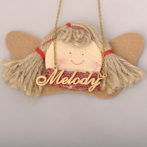 Op de afbeelding: Handgemaakte engelversiering met een lachend gezicht, blond garen haar en bruine vilten vleugels. De engel draagt een rood-wit patroon sjaal en een gouden ketting met de naam "Melody".