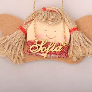May include: A gold-toned metal necklace featuring a cursive nameplate that reads Sofia with a small heart detail at the end. The necklace is displayed over a decorative wooden doll face ornament with tan twine hair, red yarn ties, and felt wings.