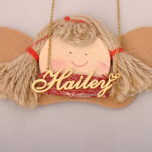 Puede incluir: Un collar dorado con el nombre "Hailey" en cursiva. El collar se muestra sobre una figura decorativa de ángel con una cara de madera, pelo de hilo y detalles rojos. El fondo es gris claro.