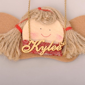 May include: A gold-coloured necklace with the name "Kylee" in a cursive font. The necklace is displayed on a decorative angel figure with a wooden face, yarn hair, and red accents.