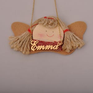 May include: A gold-colored necklace with the name "Emma" in cursive script hangs in front of a decorative angel. The angel has a wooden face, yarn hair, and felt wings. A red scarf is around the angel's neck.