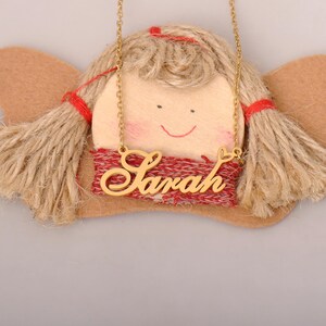 May include: A gold-toned metal necklace featuring a cursive nameplate that reads Sarah with a small heart detail. The necklace is displayed over a decorative craft ornament made of light-colored wood, tan twine hair, and red fabric accents.
