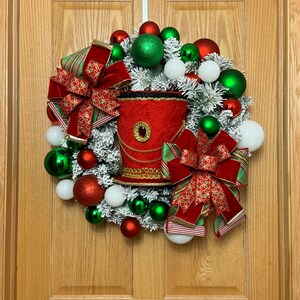 May include: A festive Christmas wreath with red, green, and white ornaments. The wreath features a red decorative hat with gold trim and two large red bows with green and gold accents. The wreath is hanging on a wooden door.