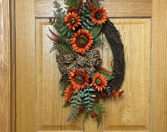 May include: A decorative oval wreath featuring orange sunflowers, green fern leaves, and a leopard print bow. The wreath is made of woven brown twigs and is hanging on a wooden door.