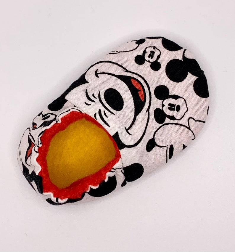 May include: A white fabric slipper with a black and red Mickey Mouse design. The slipper has a yellow felt lining.
