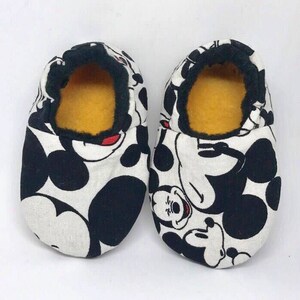 May include: A pair of white and black Mickey Mouse slippers with a yellow fleece lining. The slippers are made of fabric and have a black trim around the opening.