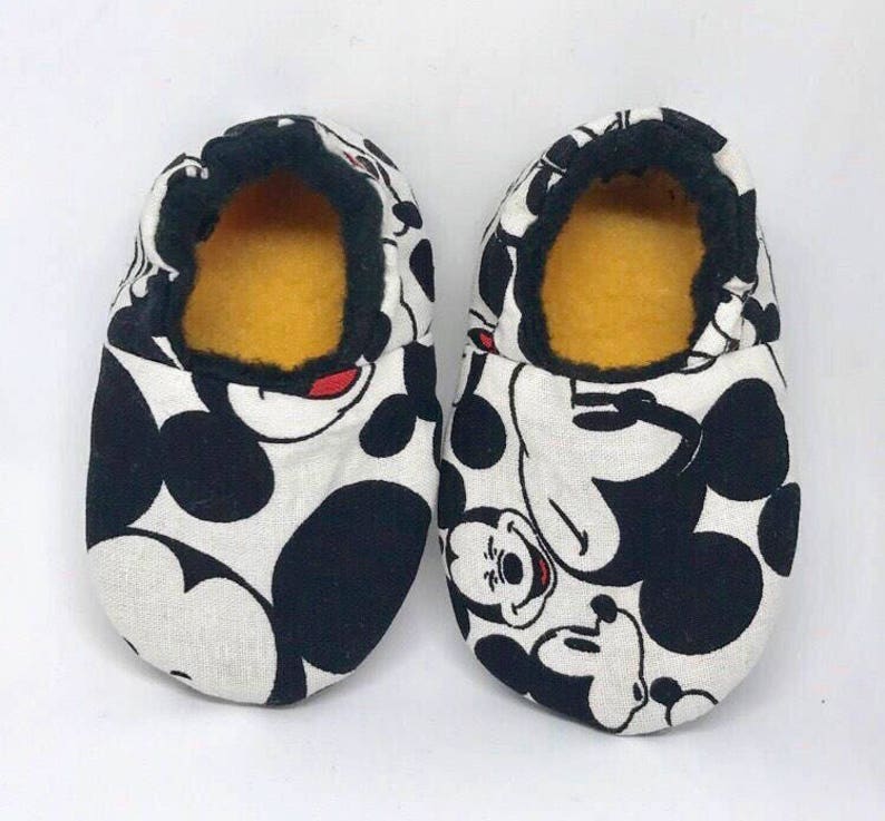 May include: A pair of white and black Mickey Mouse slippers with a yellow fleece lining. The slippers are made of fabric and have a black trim around the opening.