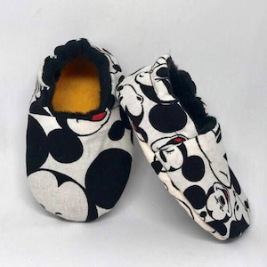 May include: A pair of white baby slippers with a black and red Mickey Mouse print. The slippers have a black fuzzy lining.