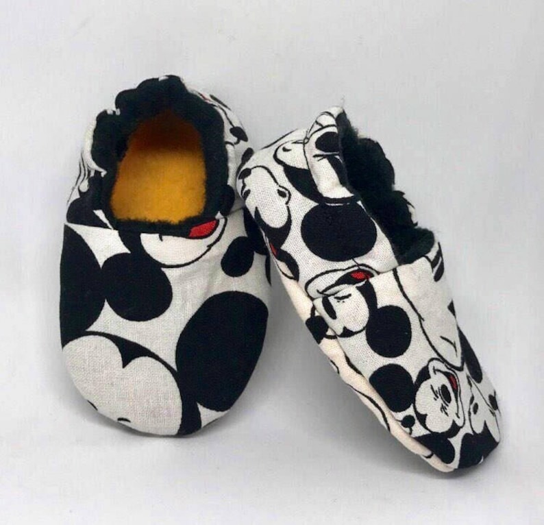 May include: A pair of white baby slippers with a black and red Mickey Mouse print. The slippers have a black fuzzy lining.