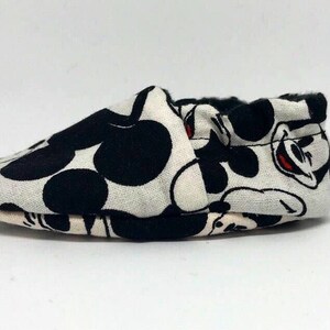 May include: A pair of white baby shoes with a black and white Mickey Mouse print.