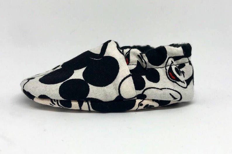 May include: A pair of white baby shoes with a black and white Mickey Mouse print.