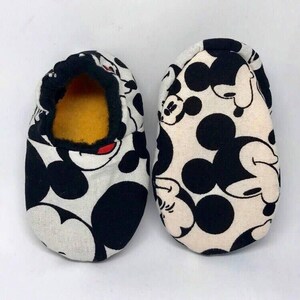 May include: A pair of white baby slippers with a black and white Mickey Mouse print. The slippers have a black trim and a yellow lining.