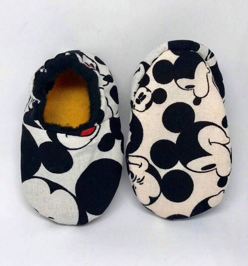 May include: A pair of white baby slippers with a black and white Mickey Mouse print. The slippers have a black trim and a yellow lining.