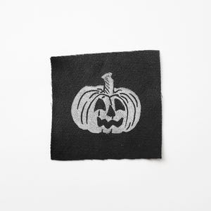 May include: A black square patch featuring a white, stylized jack-o'-lantern design. The pumpkin has a carved face with triangular eyes and a wide, toothy grin. The patch is likely for apparel or crafting.