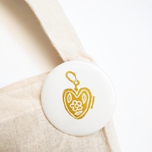 May include: A white button with a gold heart-shaped locket design, featuring a flower inside. The button is attached to a beige canvas tote bag. The locket design is detailed and elegant.