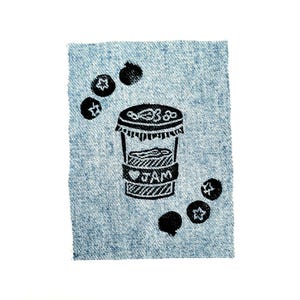 May include: A black and white linocut print of a jam jar with the word "JAM" on a denim background. The print is surrounded by stylized blueberries. The denim is a light blue color.