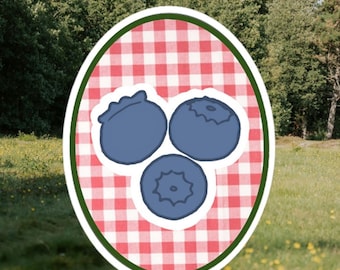 May include: An oval sticker with three blue blueberries on a red and white gingham pattern. The sticker has a green border. The background shows a grassy field and trees.