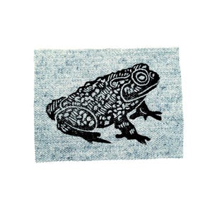 May include: A black and white illustration of a frog on a square of light blue fabric. The frog is detailed with intricate patterns and textures, giving it a unique, artistic appearance. The fabric has a subtle, textured appearance.