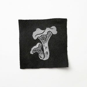 Puede incluir: Un parche de tela negra cuadrada con una ilustración en gris claro de dos setas de rebozuelo. Las setas están detalladas con branquias y tallos visibles. El parche es para coser o planchar sobre ropa o accesorios.