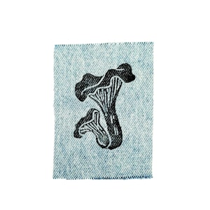 May include: A black and white mushroom graphic is printed on a small, square piece of light blue denim fabric. The illustration depicts two mushrooms with detailed gills and textured caps. The denim has a subtle, woven pattern.