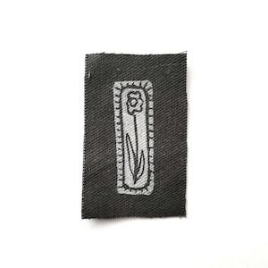 May include: A small, rectangular black fabric patch with a white floral design. The design features a stylized flower with a stem and leaves, enclosed within a white rectangular border. The fabric has a textured appearance.