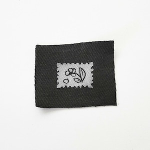 Può includere: Una piccola toppa quadrata di tessuto nero con un disegno bianco in stile francobollo. Il francobollo presenta una semplice illustrazione di un fiore con foglie e bacche. La toppa è probabilmente per cucire o creare.